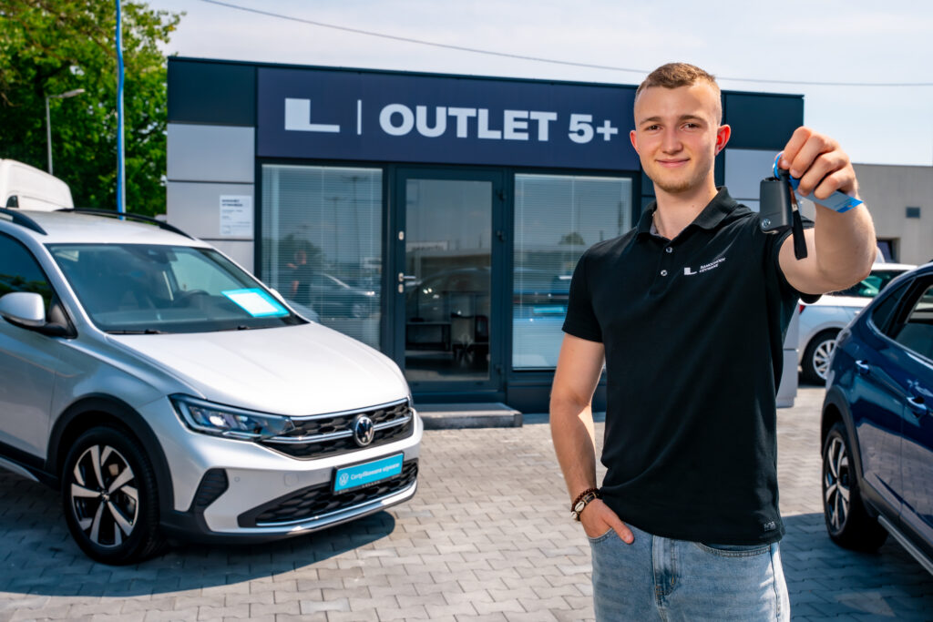 Asystent sprzedaży wyciągający dłoń z kluczykiem samochodowym w stronę klienta. W tle samochód marki Volskwagen oraz salon Outlet 5+ LELLEK Opole